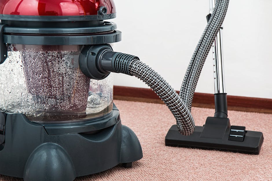 A professional cleaner wearing a white protective suit, face mask, and gloves is performing a deep cleaning or sanitisation of a beige carpet in a bright, modern living room. The room features a light